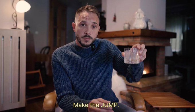 A man in a blue sweater sitting by a fireplace holding a glass of a light-colored beverage, with the subtitle 'Make the JUMP.'