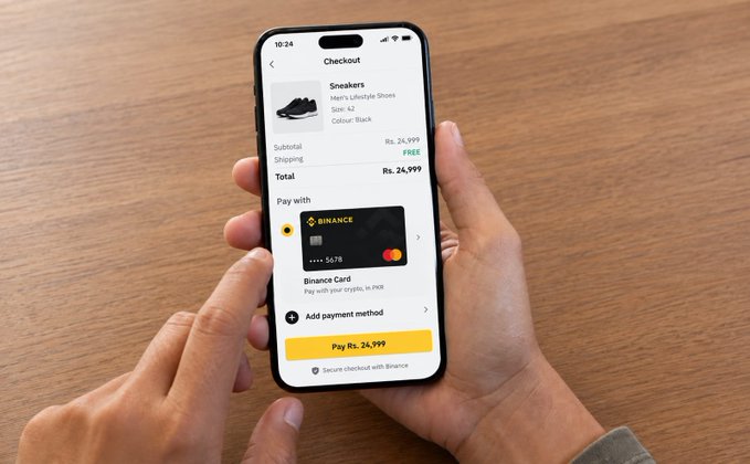 A person holds a smartphone displaying a checkout screen for a black sneaker order totaling Rs. 24,999, with payment options including Binance Card and 'Pay with crypto, in Fiat'.