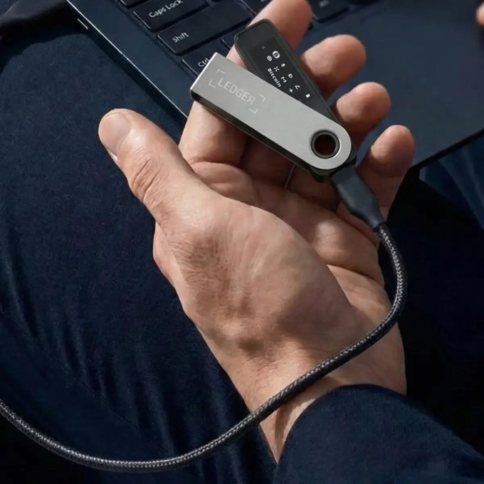 A close-up of a hand holding a silver Ledger hardware cryptocurrency wallet connected via a braided USB cable, with a laptop keyboard visible in the background.