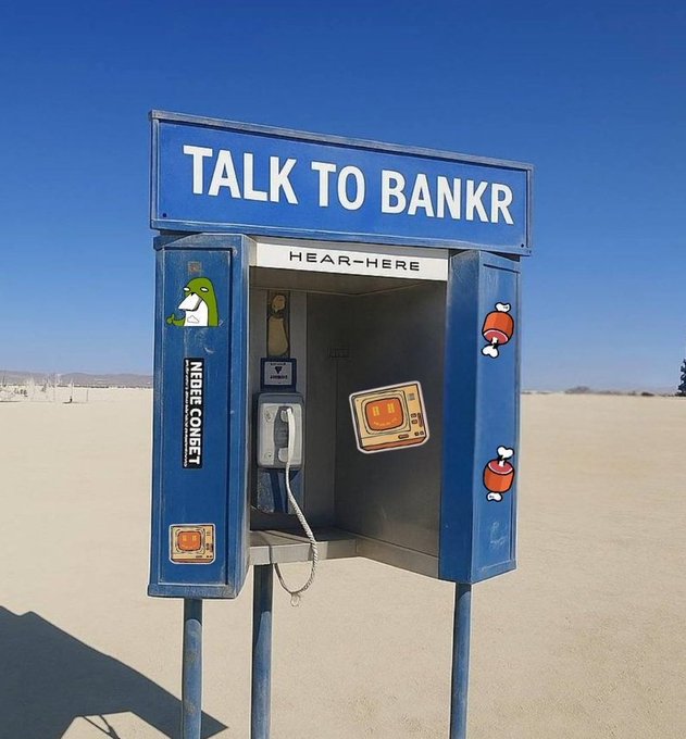 A blue phone booth in a desert landscape modified to read 'TALK TO BANKR' and 'HEAR-HERE' with 'NEEDECOMET' on the side, decorated with cartoon stickers.