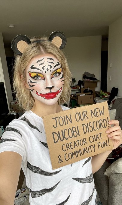 A woman in tiger face paint wearing bear ears and a tiger-striped shirt holds a cardboard sign reading 'JOIN OUR NEW DUCOBI DISCORD! CREATOR CHAT & COMMUNITY FUN!'