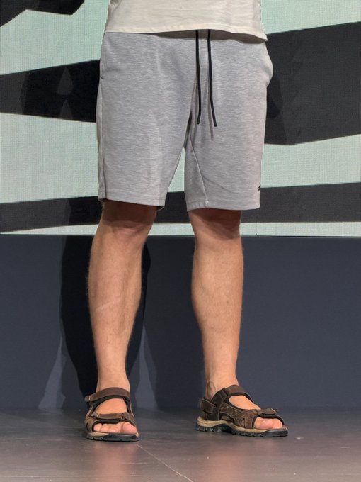 A photo showing a person's lower half wearing gray drawstring shorts and brown Teva-style sandals, standing on a stage with a digital screen in the background.