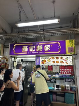 Customers line up at a noodle stall featuring a bright yellow and blue sign with Chinese characters reading '基記麵家' and 'JI KEE NOODLE HOUSE' along with a 'Michelin' recommendation sticker. The stall displays various menu items on the wall behind the counter.