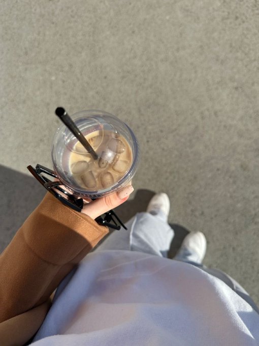 A first-person view shows a hand holding an iced coffee drink with a straw, wearing a brown long-sleeved top, with white pants and shoes visible below on a concrete surface.