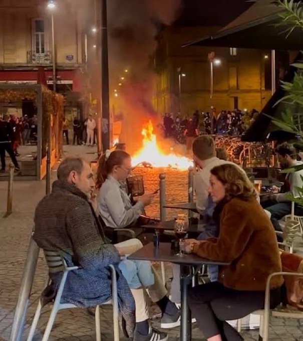 A group of people sit calmly at an outdoor café, enjoying drinks while a large fire rages in the street behind them and crowds gather in the background. The image appears to depict recent unrest in France, which the post contrasts with El Salvador by claiming the Central American country has become "verifiably safer" than France and 160 other nations.