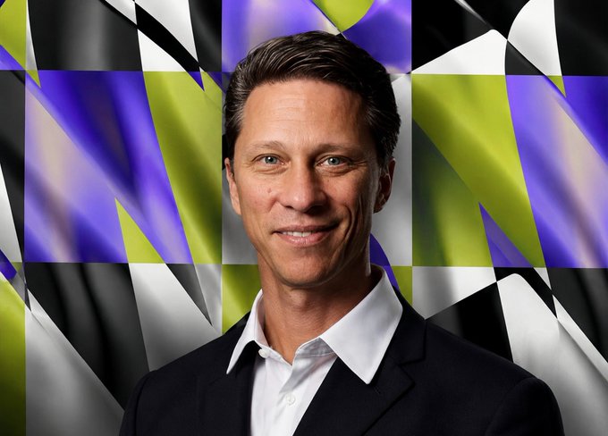 A professional headshot of a smiling man in a black suit and white shirt, set against a colorful abstract background of purple, yellow, and black geometric shapes.