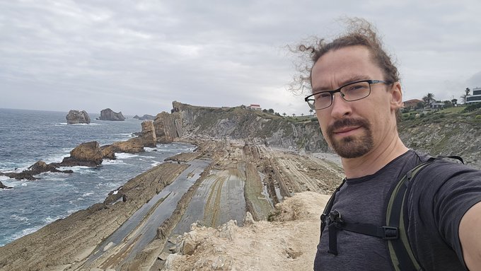 A man with glasses and a backpack takes a selfie on a coastal cliff, with rocky shorelines, ocean, and a cloudy sky stretching into the distance.