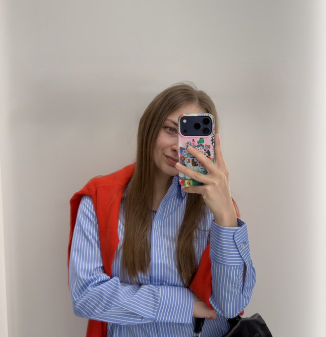 A woman takes a mirror selfie in a blue striped shirt and red cardigan, holding a colorful phone case, while sharing her crypto portfolio history—from her first XRP purchase to her favorite Ethereum.