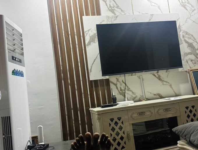 The image shows a person's feet resting on a table with their legs crossed over each other. They are wearing socks that have a unique design featuring a diamond pattern. The person appears to be sitting or standing near a television, which is turned off. In the background, there is a white air conditioner and a wooden wall.