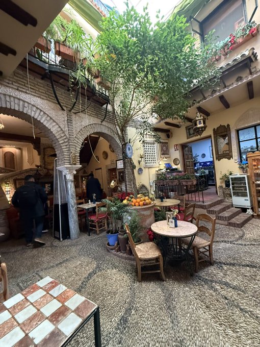 A traditional Mediterranean-style courtyard café features stone archways, lush greenery, and small tables adorned with fresh oranges, where patrons gather for a morning coffee. The serene, sunlit setting reflects the relaxed energy of a "Gm everyone" greeting, capturing a peaceful start to the day in a charming, rustic atmosphere.
