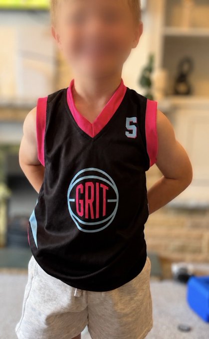 A young child with a blurred face stands with hands on hips wearing a black and red sleeveless jersey with the number '5' and a circular 'GRIT' logo on the front, paired with light gray shorts.