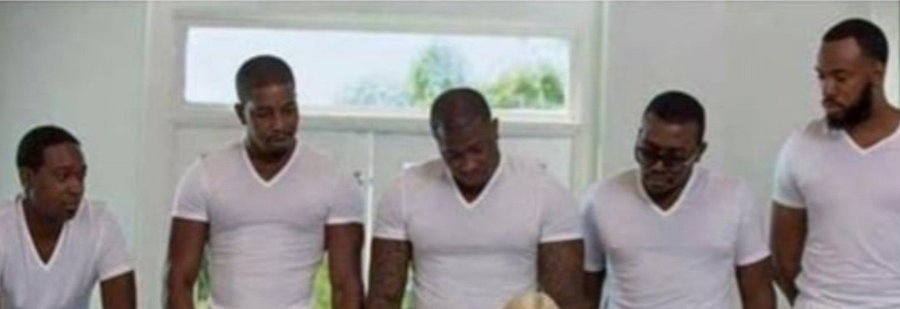Five men wearing matching white v-neck t-shirts stand shoulder-to-shoulder in front of a window with white trim, all looking downward in a contemplative or prayerful pose that suggests unity and anticipation, fitting the teaser caption "It's coming…." as a promotional hint at an upcoming announcement, collaboration, or release.