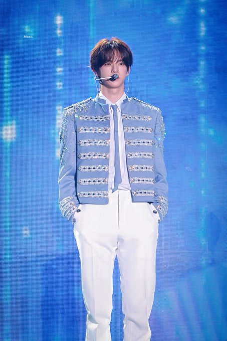 A performer wearing a sparkling light blue military-style jacket and white pants stands on stage against a blue backdrop, wearing a headset microphone.