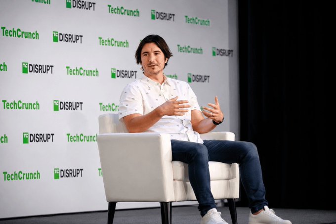 Vlad Tenev, CEO of Robinhood, speaks on stage at TechCrunch Disrupt, seated in a white armchair against a backdrop featuring repeated "TechCrunch Disrupt" logos in green and black.