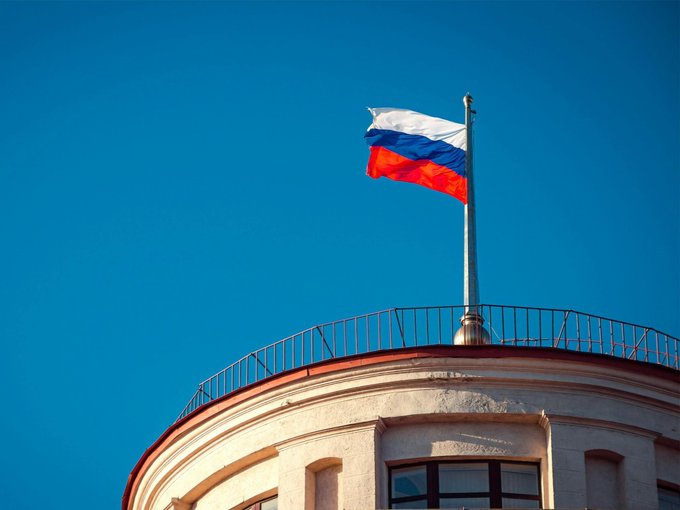 The Russian tricolor flag waves atop a domed building against a clear blue sky, symbolizing the State Duma where lawmakers are set to finalize crypto regulations limiting retail investor purchases to $3,800 annually.