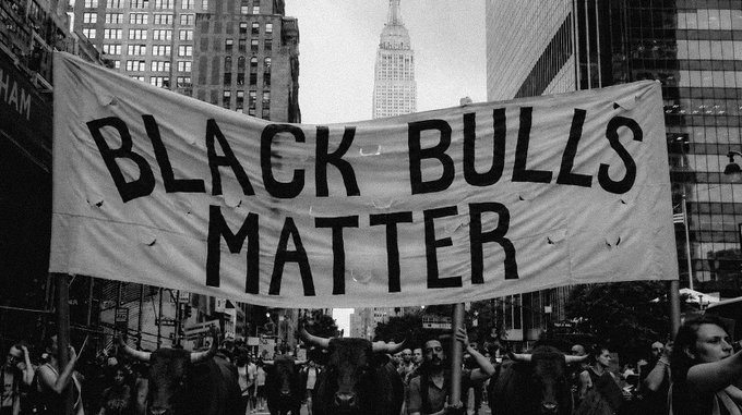 A black-and-white photograph of a protest march in a city (likely New York with the Empire State Building visible) where demonstrators carry a large banner reading 'BLACK BULLS MATTER' alongside what appear to be bull figures.