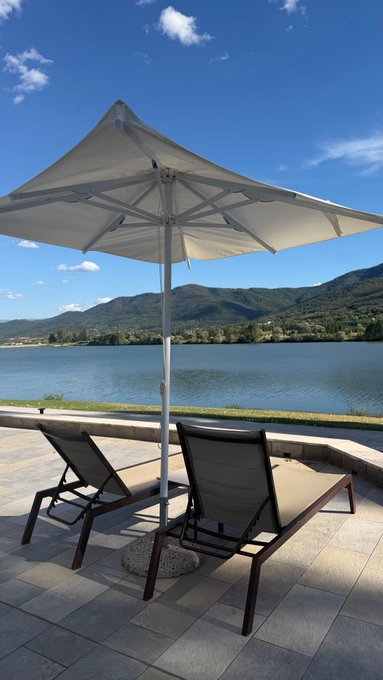 Two dark lounge chairs sit under a large beige umbrella on a tiled patio overlooking a serene blue lake surrounded by green mountains under a clear sky.