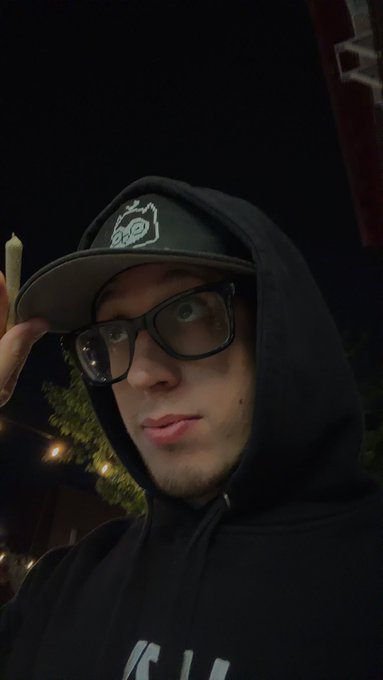 A young man wearing glasses and a black hoodie and cap takes a selfie at night, with string lights visible in the background.