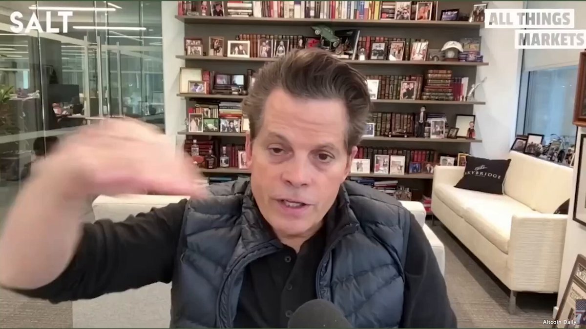 The image features a man wearing a black vest, sitting in front of a bookshelf filled with numerous books. The man appears to be talking about Bitcoin or cryptocurrency, as indicated by his hand gesture towards the camera. The background includes a couch and a window, suggesting that he is in an office setting. The caption below the image reads "Anthony Scaramucci: Bitcoin is bottoming. Now is the time to buy. Here is why #bitcoin". This implies that Scaramucci is discussing the current state of Bitcoin or cryptocurrency markets and making a case for investing in it at this time.