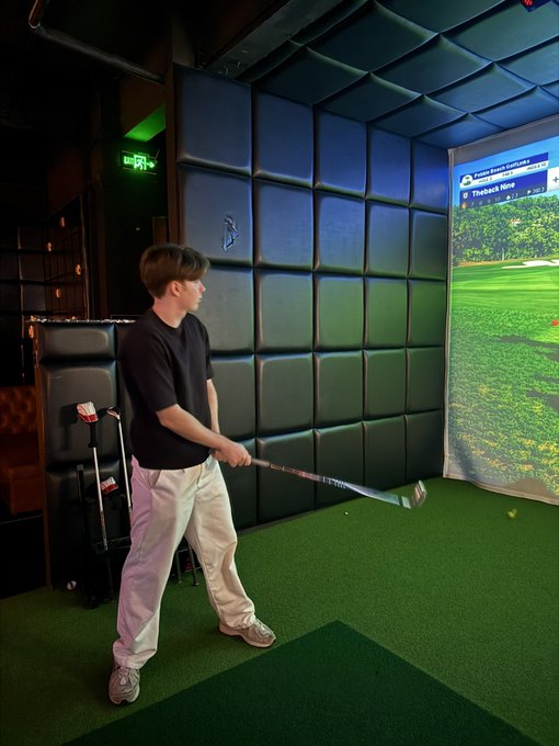 A young man in a black shirt and white pants practicing golf swings in an indoor golf simulator with 'Pebble Beach Golf Links' and 'Thalback Nine' displayed on the screen.