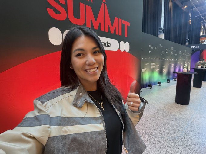 A woman with long dark hair takes a selfie at what appears to be the "Summit Canada" event, smiling broadly and giving a thumbs up in front of a red and black backdrop with "SUMMIT" and "ada" visible, along with a "STAGE" sign in the background, marking the conclusion of the conference.