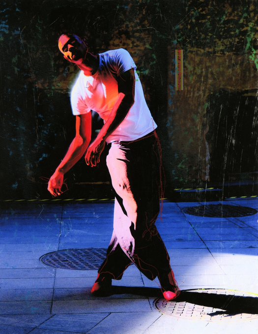 A stylized, high-contrast photograph shows a man in a white t-shirt and dark pants standing outdoors with rain falling around him, illuminated by dramatic red and blue lighting.