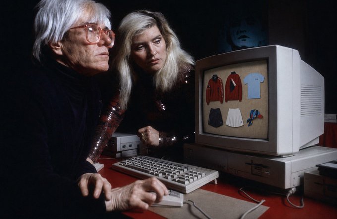 An older man with white hair and red glasses sits at a vintage Apple computer with a younger blonde woman; the monitor displays images of clothing items.
