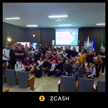 The image features a large group of people posing for a photo in front of a projector screen displaying a presentation slide with the text "Zcash". The attendees are seated in chairs, some standing while others sit down. There are at least 14 individuals visible in the picture, all gathered together and seemingly engaged in conversation or enjoying the event. The presence of a projector screen suggests that this gathering might be related to an educational or professional event where participants discuss topics such as technology, privacy, and innovation.