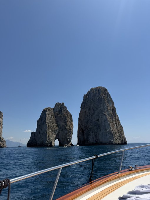Two large rock formations, a cliff and a cave, dominate the horizon of a deep blue sea, with a boat visible on the water's surface.