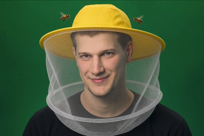 A man wearing a yellow beekeeper's hat with mesh netting and two bees flying on either side of his head, set against a green background.