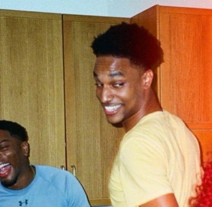 Two young men share a lighthearted exchange in what appears to be a kitchen with wooden cabinets; one in a tan t-shirt smiles at the camera while another in a gray shirt laughs in the background.