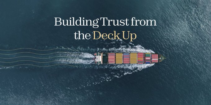 An aerial photograph of a cargo ship loaded with colorful containers sailing across dark blue ocean water, overlaid with white and gold text reading 'Building Trust from the Deck Up'.