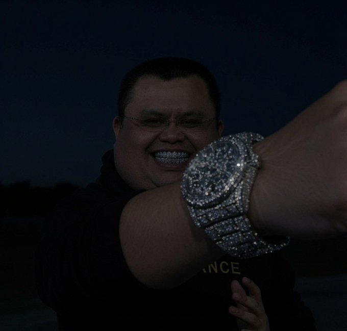 A man wearing glasses and a black shirt smiles while showing off a diamond-encrusted "CZ" grillz and a large diamond-encrusted watch, with partial text "NCE" visible on his shirt—visually celebrating the post's reference to profiting from $CZ at its lows.