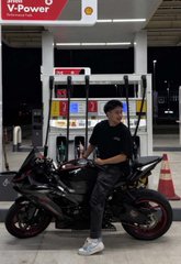 A person wearing a black shirt, cap, and face mask sits on a black and red sport motorcycle at a brightly lit V-Power Shell gas station at night.