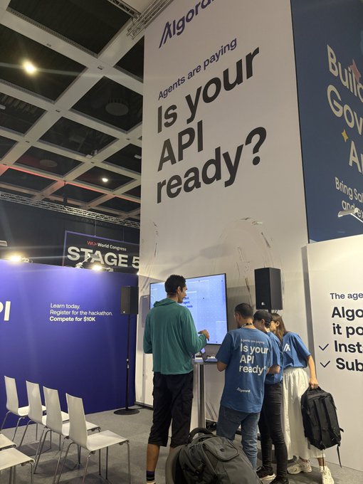 Photo from a blockchain conference showing the Algorand booth with signage reading 'Agents are paying: Is your API ready?' as attendees interact with displays.