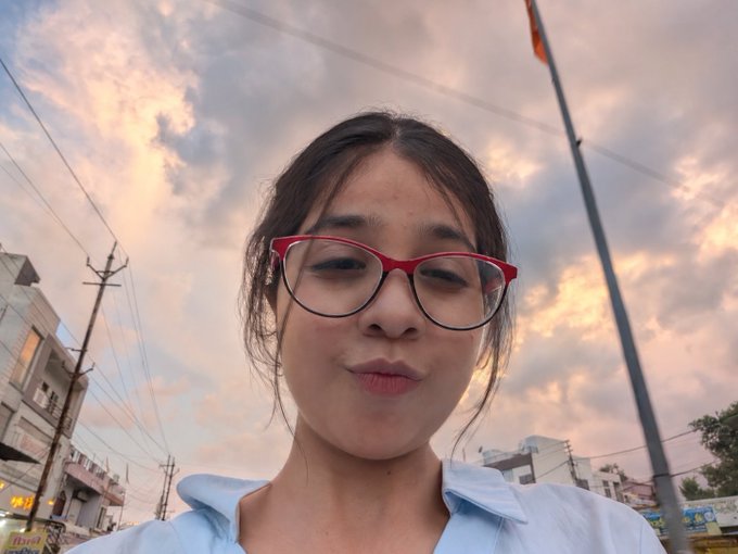 A selfie of a young woman wearing red glasses with a sunset sky and urban buildings visible in the background.
