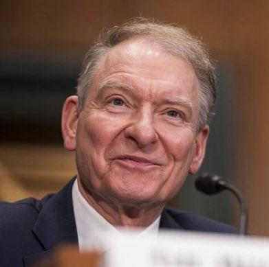 An older man in a dark suit sits at what appears to be a congressional hearing, looking forward with a slight smile.