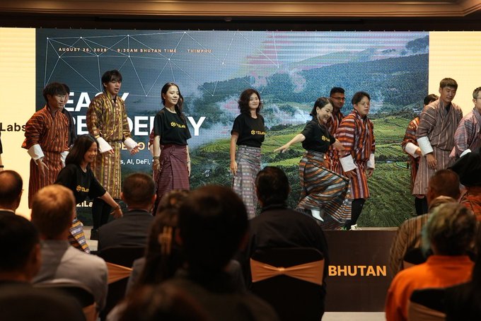A group of people wearing traditional Bhutanese striped garments stand on a stage performing for an audience; a banner behind them reads 'AUGUST 10, 2025' and '9:15AM BHUTAN TIME - THIMPHU' with the word 'BHUTAN' visible on a chair.