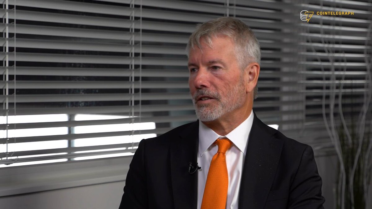 The image features an older man wearing a suit and tie with an orange tie, sitting at a desk in front of a window. The background shows a white wall with blinds covering the window. There are also some plants visible near the window. The image is related to a post on social media that discusses the potential recovery of Bitcoin after the recent crash.