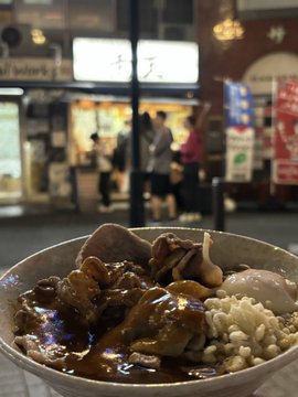 The image shows a bowl of food placed on a table, with a blurred background that includes people walking by outside a store or restaurant. The focus of the photo is on the bowl of food, which appears to be a delicious-looking meal consisting of meat and rice.