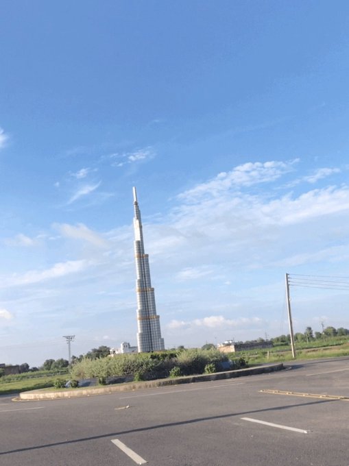 The image features a tall, white tower that resembles the Burj Khalifa, an iconic skyscraper located in Dubai, United Arab Emirates. The tower stands out against the backdrop of a clear blue sky with some clouds scattered across it. In the foreground, there is a parking lot and a street sign, adding to the urban setting surrounding the impressive structure.