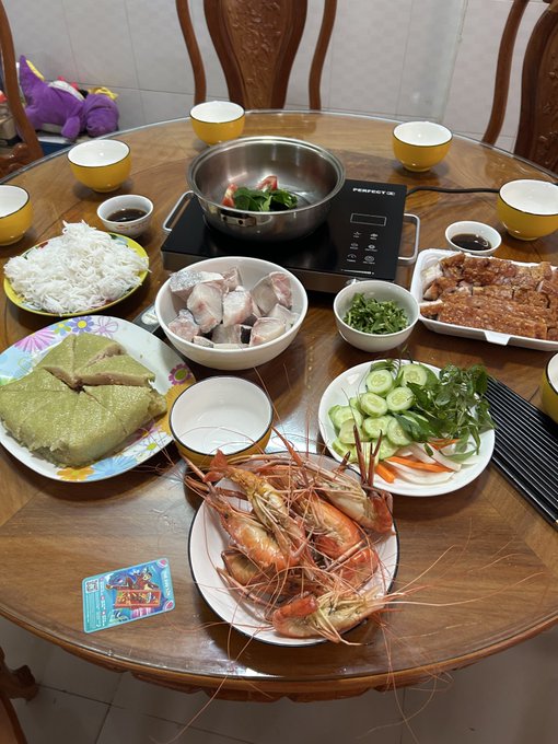 The image features a wooden dining table with various dishes of food, including shrimp, rice, vegetables, and other items. The table has several bowls containing different types of food, as well as cups and plates filled with more food. There are also chopsticks on the table, suggesting that this meal is meant to be enjoyed using these utensils. A chair can be seen in front of the table, indicating a place for someone to sit down and enjoy their meal. The post about Bitcoin cứ lòng vòng mà xuống cũng được mà xuống có thúy là nhân, which is a reference to the cryptocurrency Bitcoin.