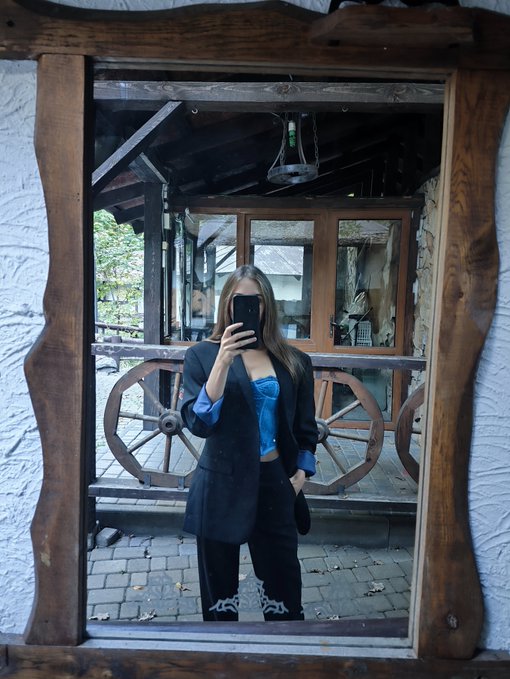 A mirror selfie of a woman wearing a black blazer and blue top, reflected in a wooden-framed mirror in what appears to be a rustic setting with wagon wheels.