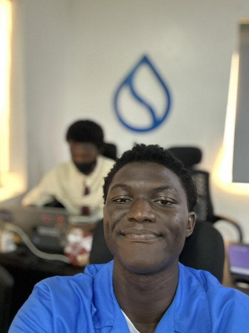 A selfie of a young man in a blue shirt smiling in an office setting, with a colleague working on a laptop in the background and a blue Sui logo visible on the wall.