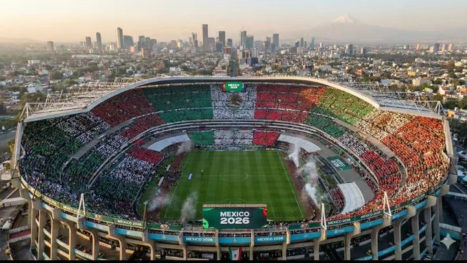 The image shows a large stadium filled with people, possibly attending a soccer game or event. The stadium has a unique design featuring a flag-like pattern painted across its roof. The colors of the flag are red, white, green, and blue. The stadium is located in Mexico City, which can be seen in the background as well. The image also includes text that reads "6月波动大", "继续震荡 关注世界杯", "6月12开赛", and "spcx上市也是在6月12，到时候看看吸行好都不拉盘的山寨别指望这时候拉也是跑路". The image captures the atmosphere and excitement of a large gathering, possibly related to an event or sports game.