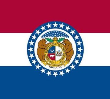 The Missouri state flag, featuring the Missouri state seal with a grizzly bear and the Latin motto "United We Stand Divided We Fall," is displayed alongside a news caption about three Missouri men charged in an alleged plot to kidnap a Bitcoin holder's parents to steal cryptocurrency. The flag's design, including the date MDCCCXX (1820) marking Missouri's statehood, serves as a visual indicator connecting the criminal incident to the state of Missouri.