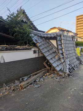 A damaged traditional-style roof structure with gray tiles collapsed onto a brick wall in an outdoor urban setting, with debris scattered on the ground.