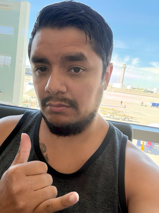 The image features a man with a beard wearing a black tank top, sitting next to a window that overlooks an airport runway. The man has his fingers crossed and is making a thumbs-up gesture. The caption of the image reads "GM #crypto Let's do the Thing!!! Party like it's 1999 fuckers!!!!! Buy the DIP". This suggests that the man might be attending or participating in a party, possibly related to cryptocurrency or digital art, with a focus on the phrase "Let's do the thing!".