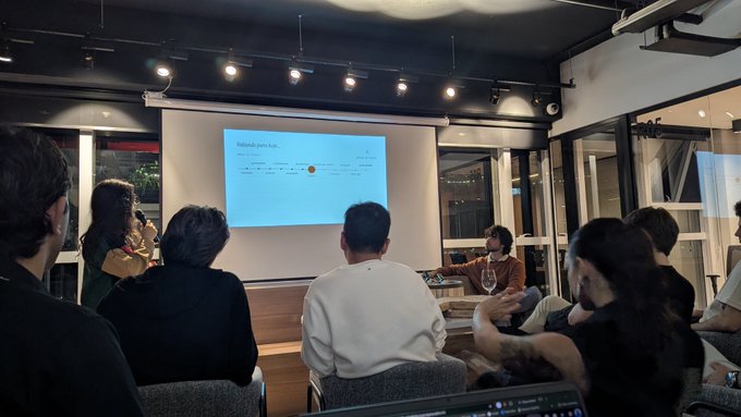 An audience sits in a dimly lit room watching a presentation on a projector screen, with one person gesturing near the screen and another holding a glass of wine.
