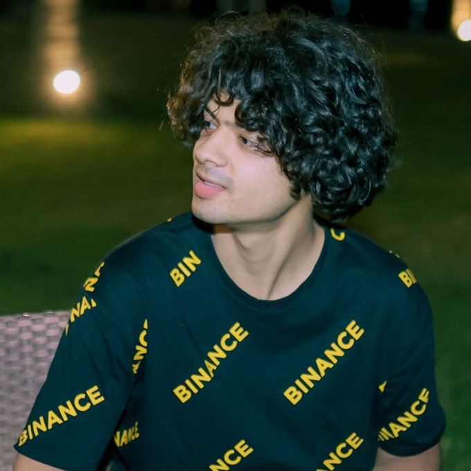 Young man with curly dark hair wearing a black t-shirt covered in yellow 'BINANCE' logos, photographed outdoors at night.